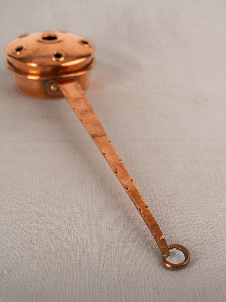 Antique French copper warming pan with long riveted handle and pierced lid, displayed on a light fabric surface.