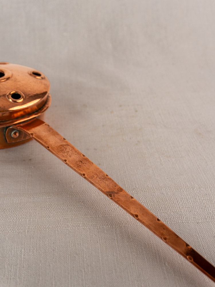 Antique French copper warming pan with vented round lid and riveted handle on light textured fabric.