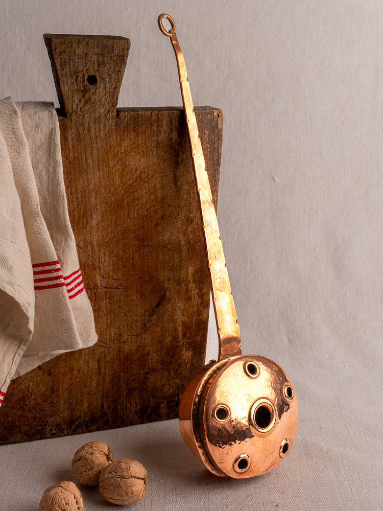 Antique French copper warming pan with riveted handle, displayed against rustic wood and a tea towel.