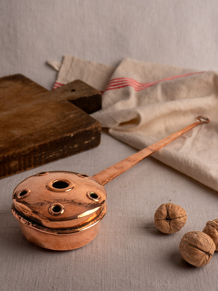 Antique French copper warming pan with lid and perforations, long riveted handle, on neutral fabric background.