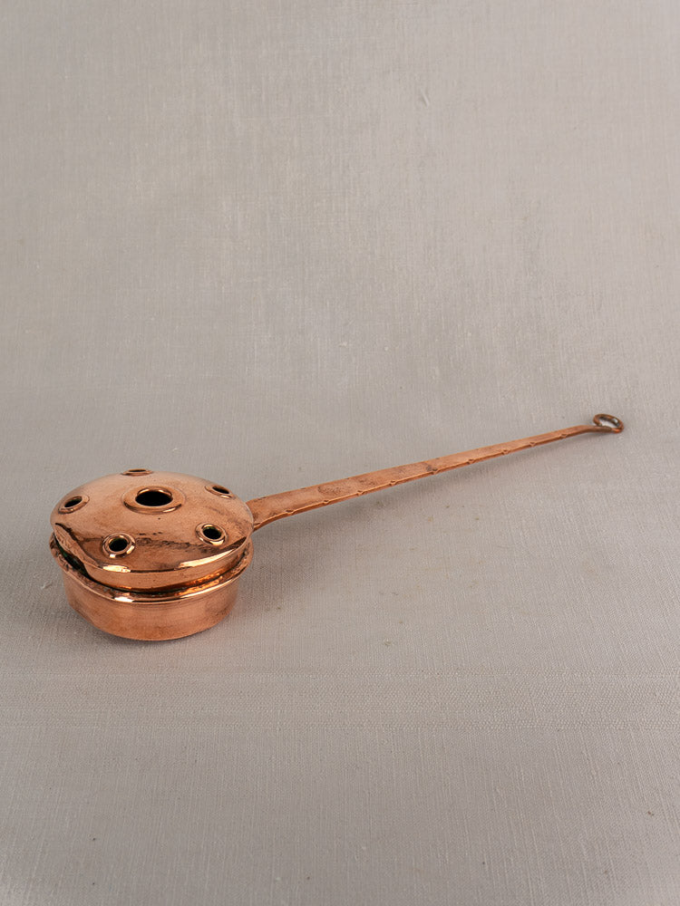 Antique French copper warming pan with lid and perforations, long riveted handle, on neutral fabric background.