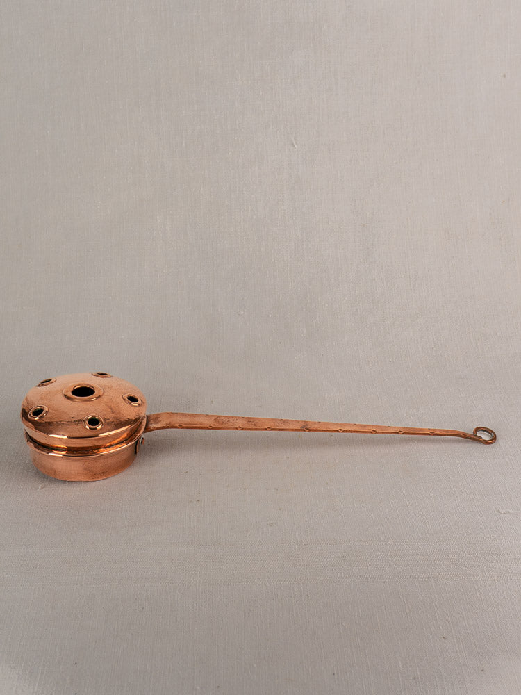 Antique French copper warming pan with long riveted handle and perforated lid, resting on neutral fabric.