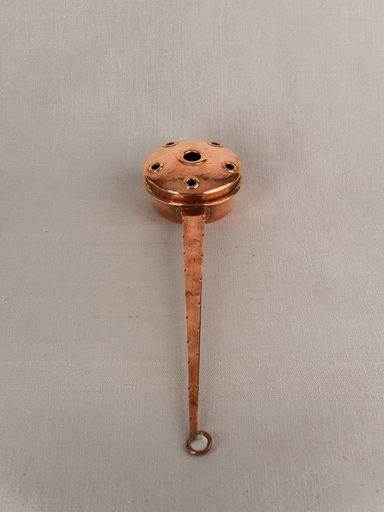 Antique French copper warming pan with round perforated lid and long riveted handle on neutral fabric background.