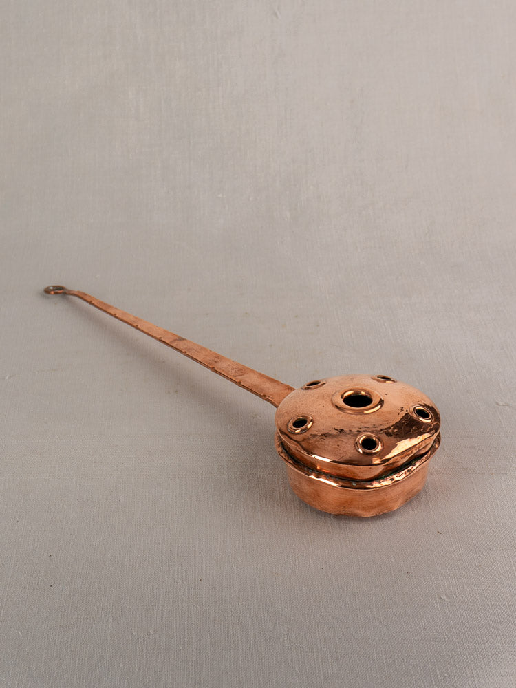 Antique French copper warming pan with long riveted handle and round vented lid on gray surface.