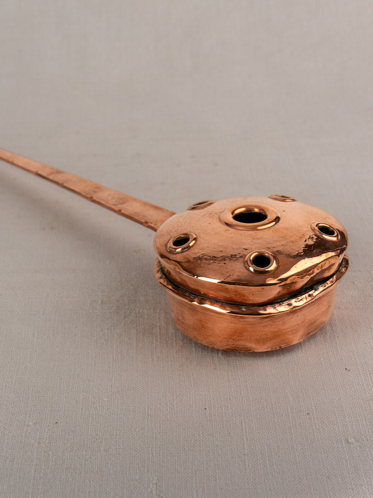Antique French copper warming pan with long riveted handle and round lidded container on neutral fabric background.
