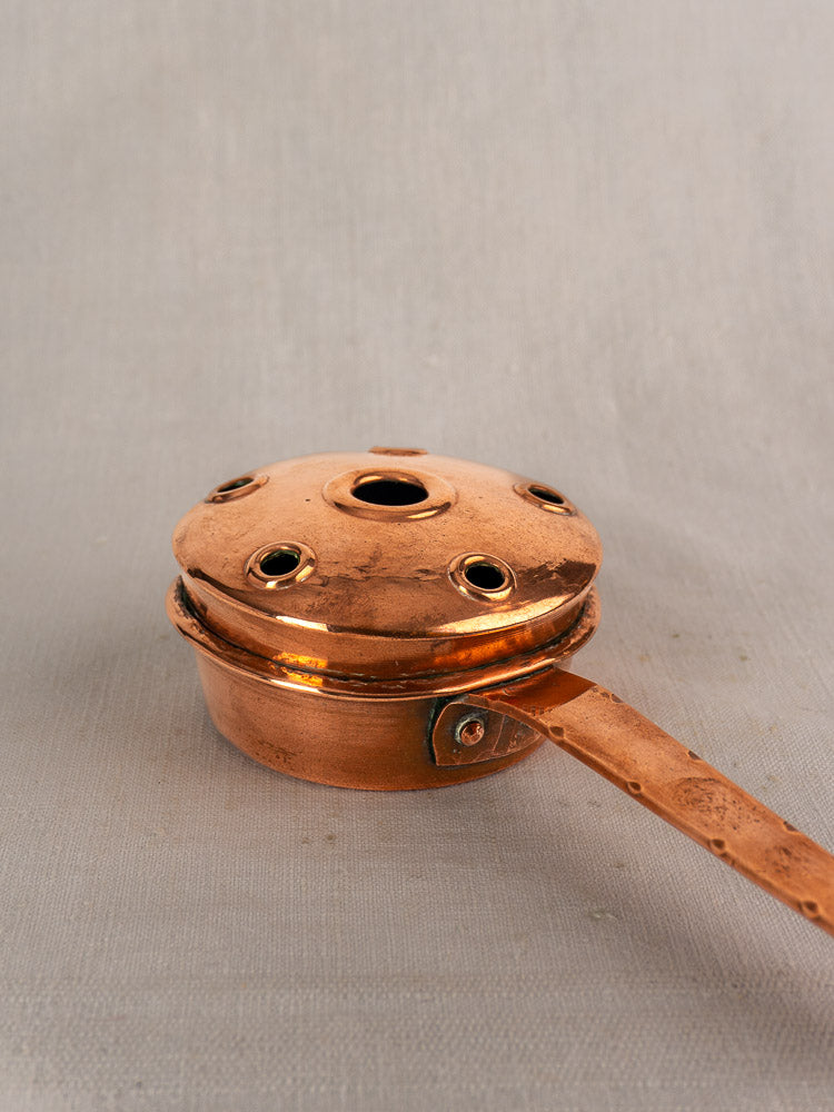 Antique French copper warming pan with round vented lid and long handle on a neutral gray fabric background.