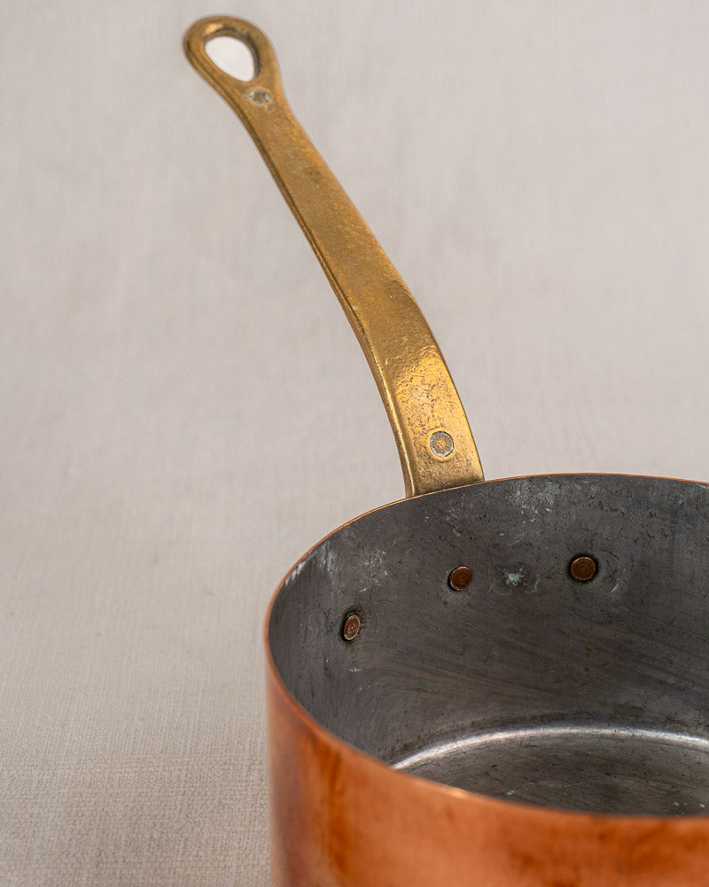 Antique French copper saucepan set of five with shiny interiors and brass handles, on a light beige surface.