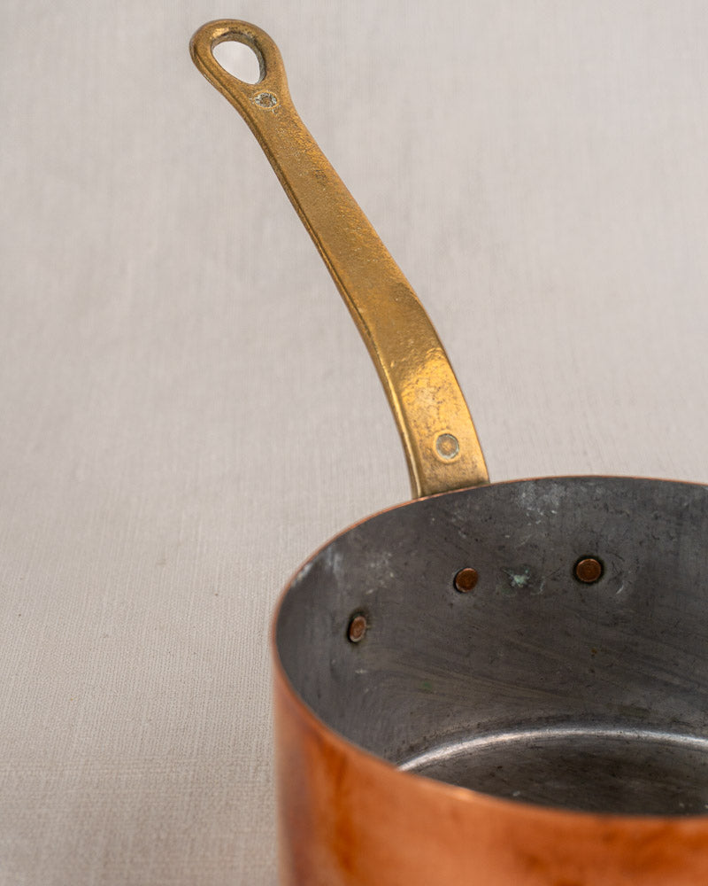 Set of five antique French copper saucepans with brass handles, tin linings, on neutral textured background.