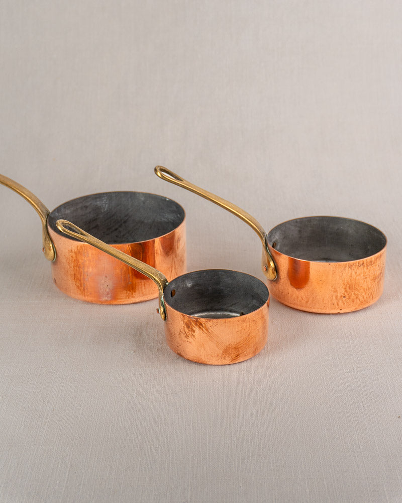 Set of five antique French copper saucepans with brass handles, varying sizes and rich patina on beige surface.