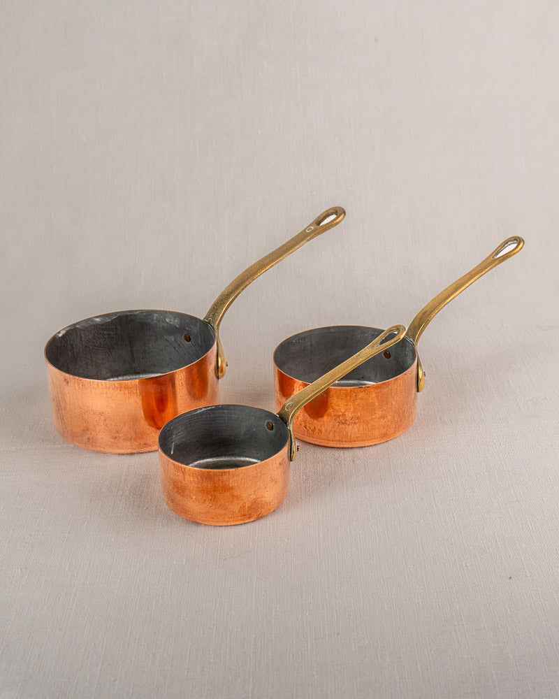 Set of five antique French copper saucepans with brass handles, nested by size on a light surface.