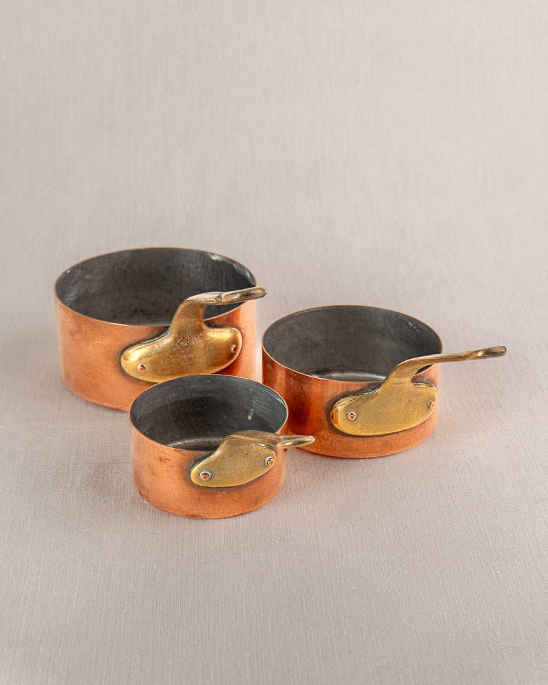 Set of five early 20th-century French antique copper saucepans with brass handles, nested on a light gray surface.