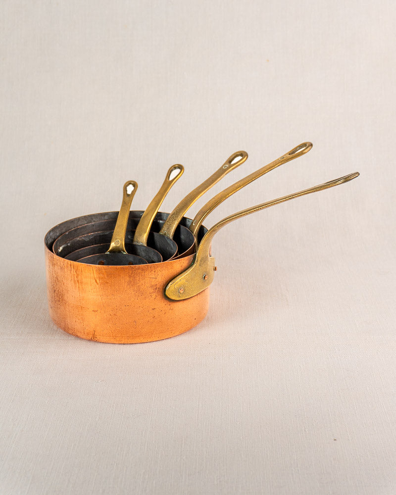 Set of five antique French copper saucepans with brass handles, nested on a light textured surface.