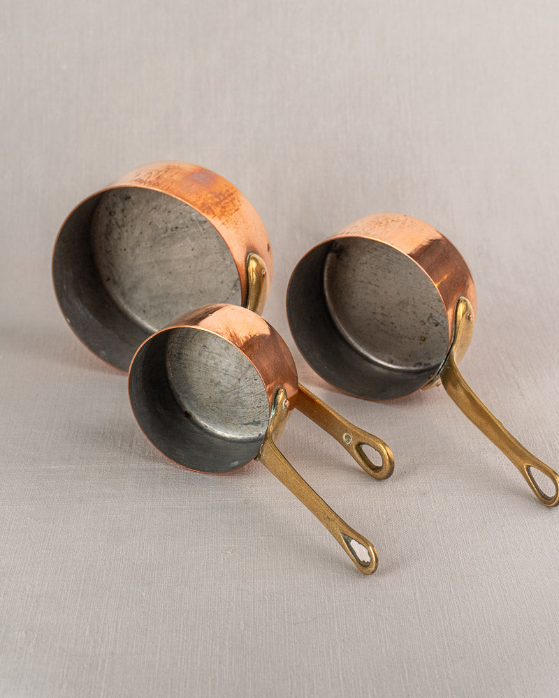 Set of five antique French copper saucepans with brass handles, patina and wear on light gray textured surface.