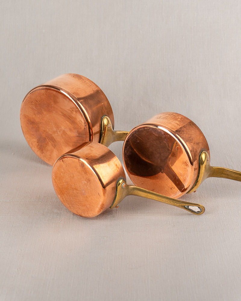 Set of five antique French copper saucepans with brass handles, polished tinned finish, on light gray surface.