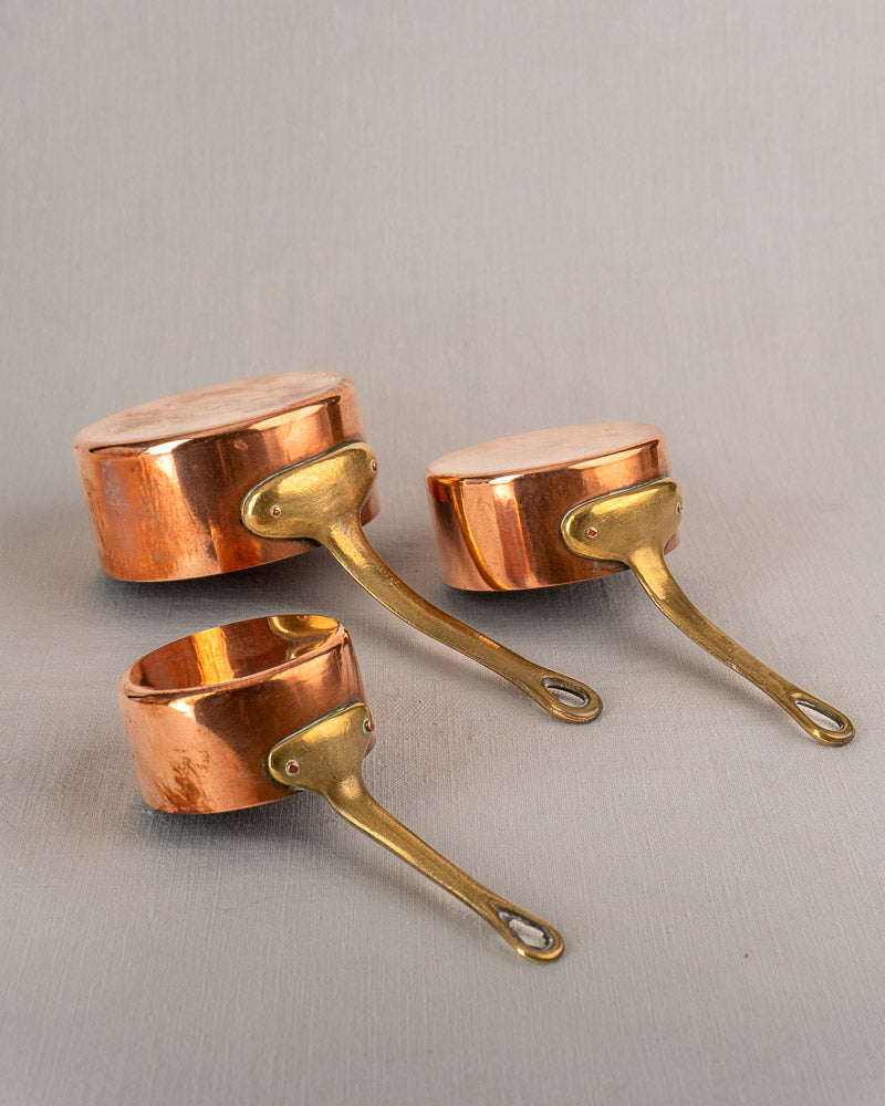 Set of five antique French copper saucepans with brass handles, graduated on a light gray textured surface.