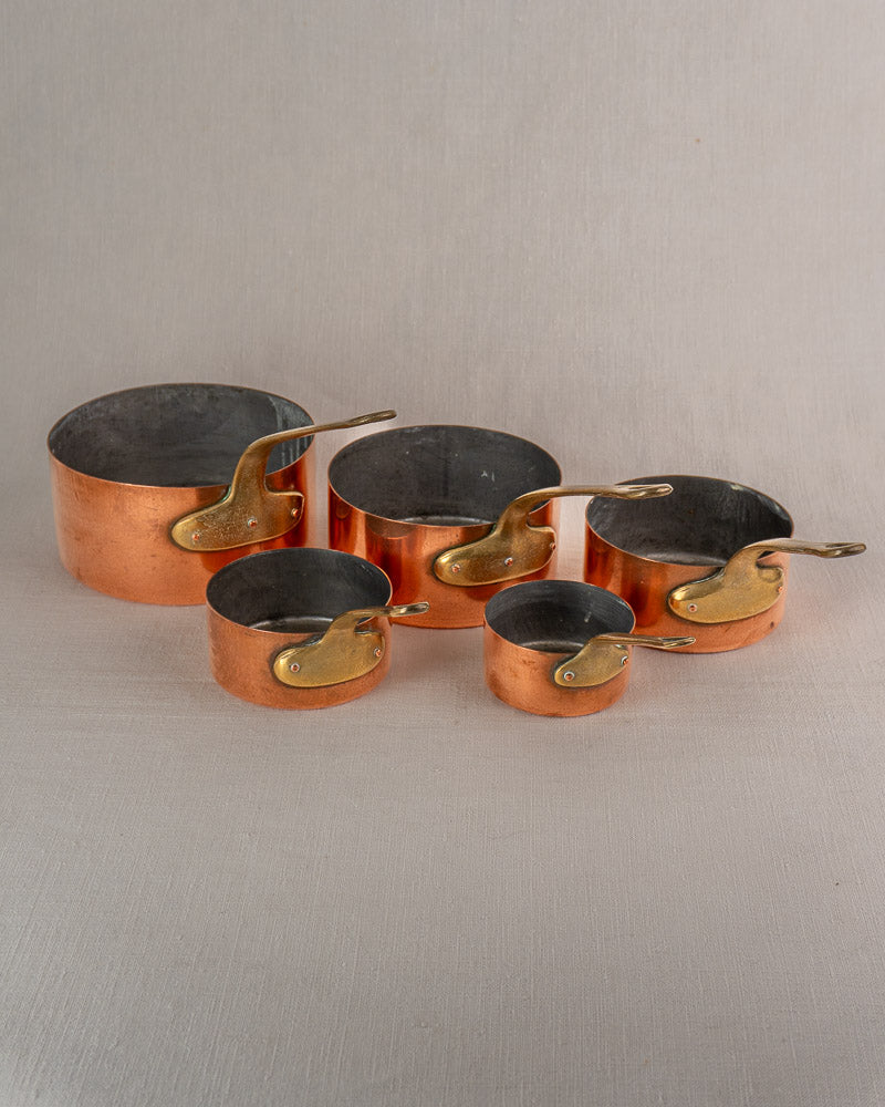 Set of five antique French copper saucepans with brass handles arranged by size on a neutral surface.