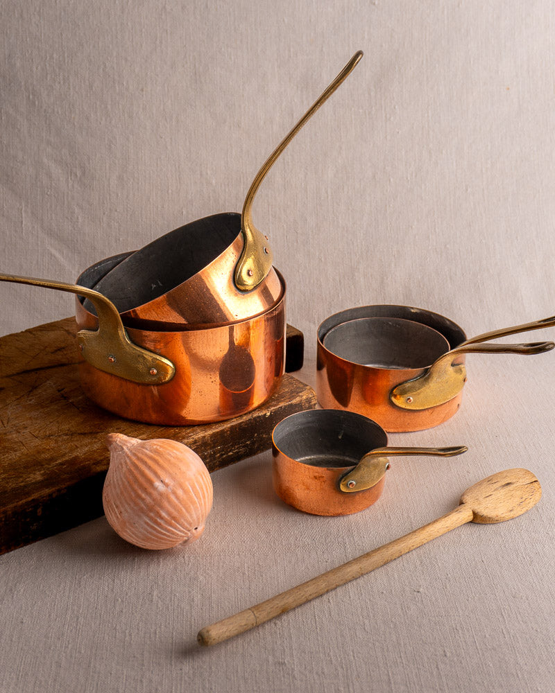Set of five antique French copper saucepans with brass handles stacked on wood, accompanied by spoon and onion.