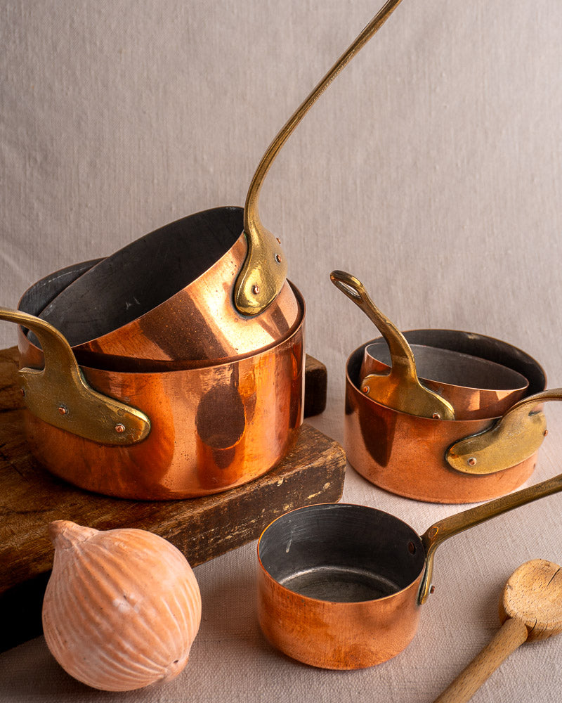 Set of five early 20th-century French copper saucepans with brass handles on wood surface, onion and spoon alongside.