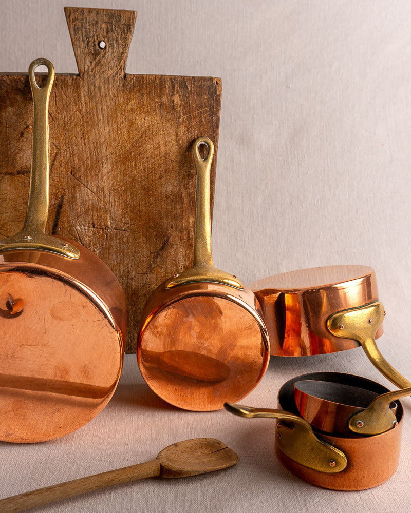 Antique French copper saucepan set of five with brass handles, shown with rustic wood board and spoon on neutral cloth.