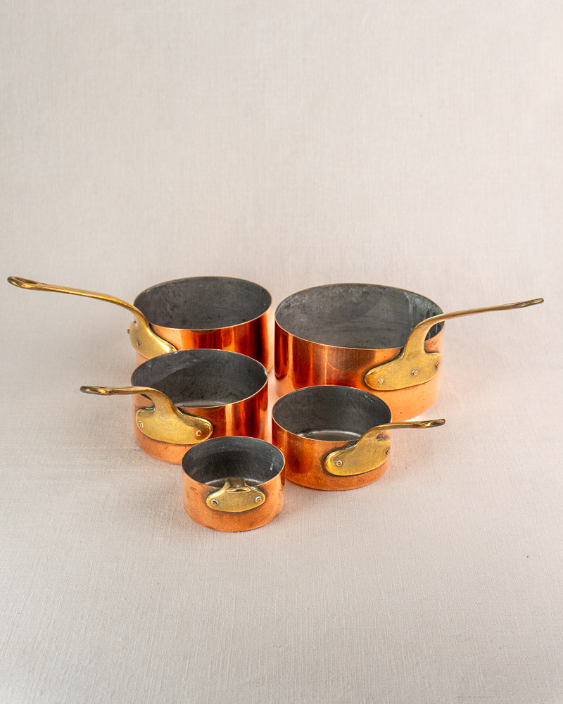 Antique French copper saucepan set of five with brass handles, tinned exteriors, dark interiors on a light surface.