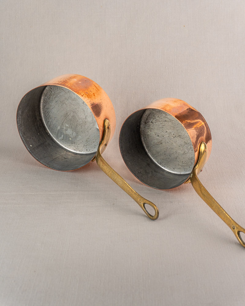 Set of five antique French copper saucepans with brass handles, aged patina and inner tarnish on light gray surface.
