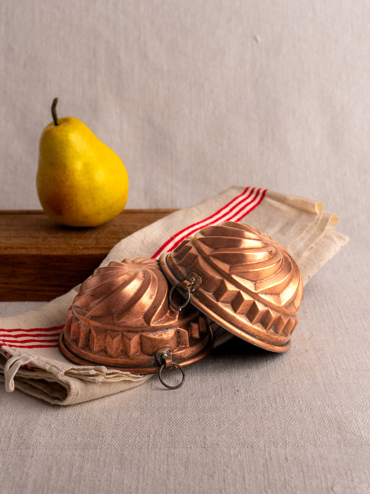 Pair of Antique French copper dessert molds with spiral relief on beige striped cloth, yellow pear and wood block behind.