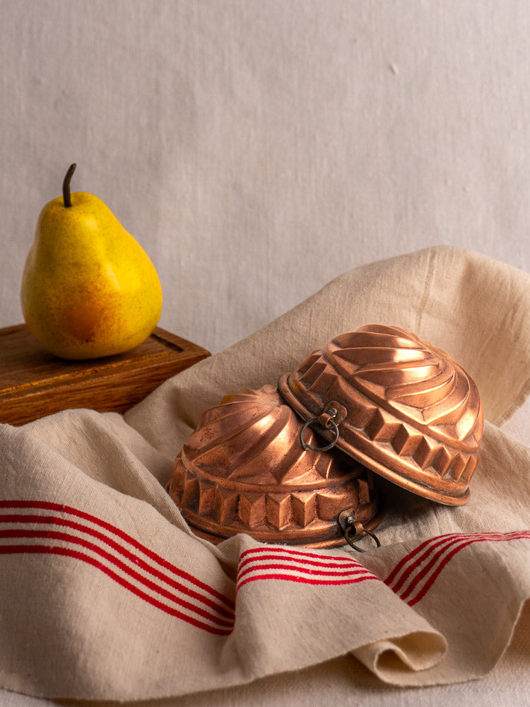 Antique French copper dessert molds with spiral relief, matched pair on striped beige cloth, yellow pear nearby.