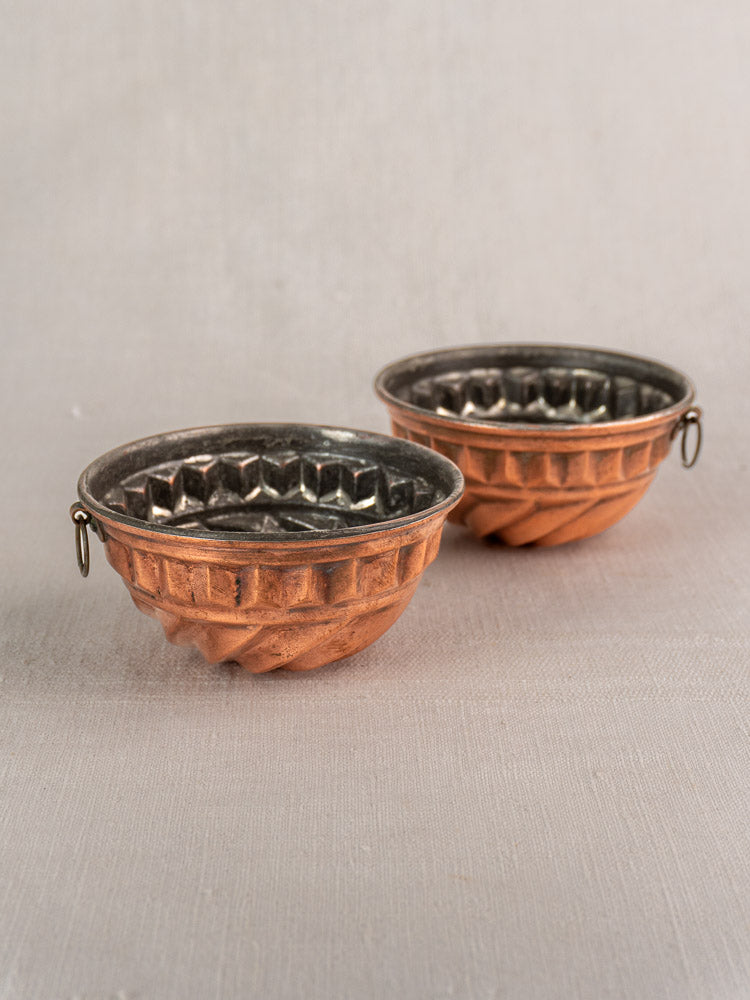Pair of antique French copper dessert molds with spiral relief on a neutral fabric surface.