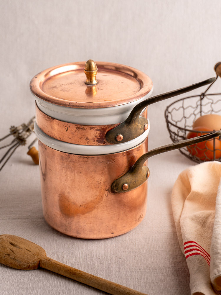 Antique French copper bain marie with Apilco porcelain insert, on cloth with spoon, whisk, eggs, and striped towel.