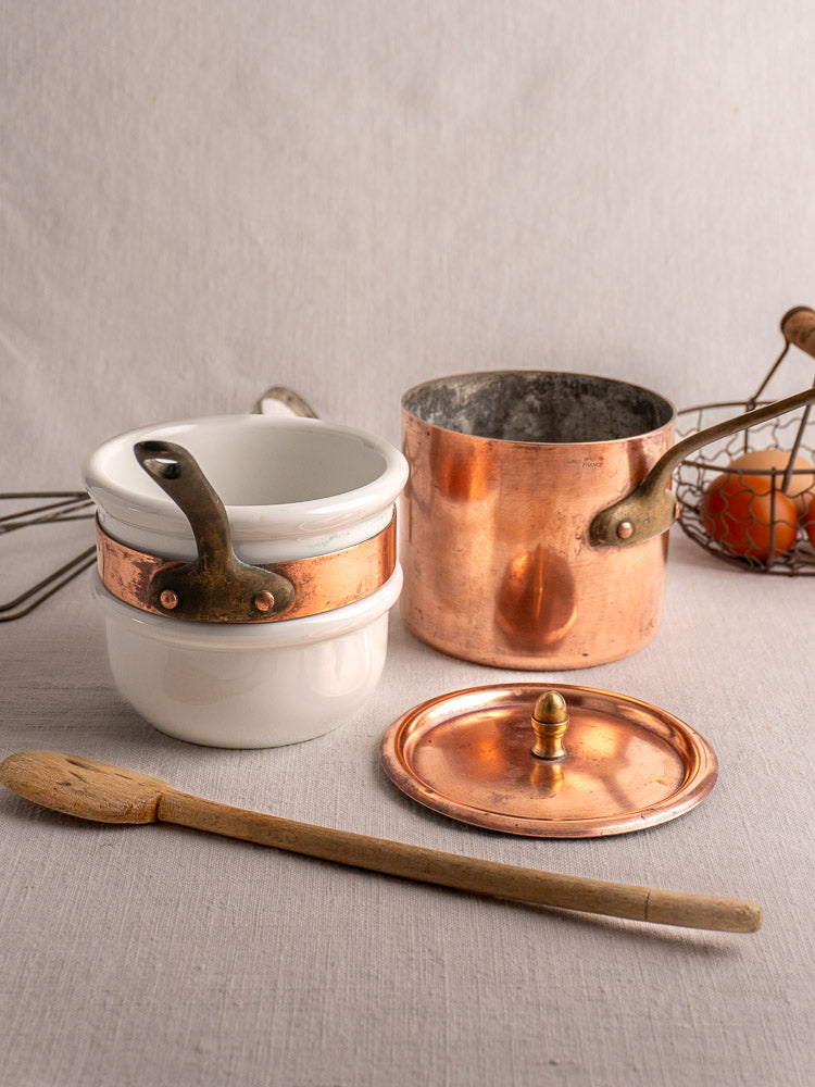 Antique French copper bain marie with Apilco porcelain insert, lid, spoon, and wire egg basket on beige fabric.