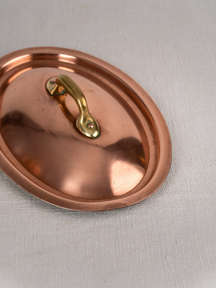 Vintage French oval copper cocotte with brass handles and domed lid on light fabric background.