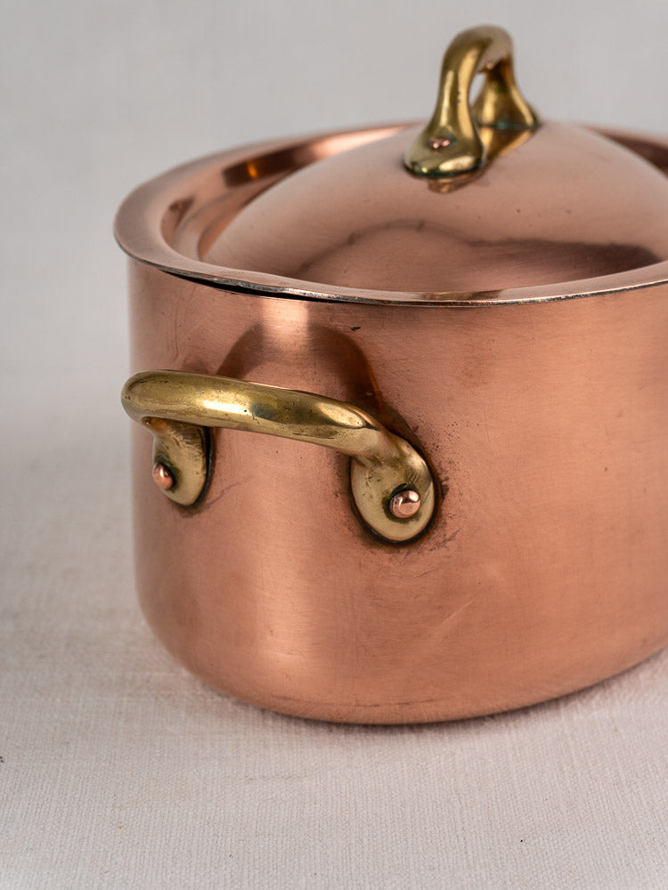 Vintage French copper cocotte with brass handles and domed lid on a light textured surface.