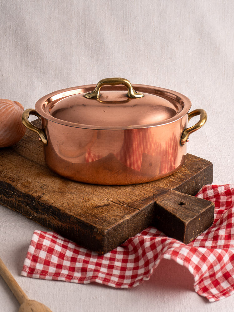 Vintage French copper cocotte with brass handles and domed lid on rustic wood board with cloth.