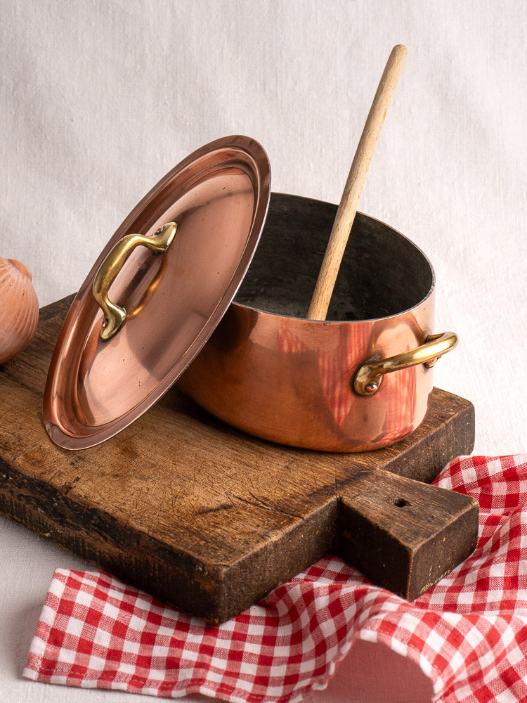 Vintage French copper cocotte with brass handles and domed lid on rustic wood board, red checkered cloth nearby.