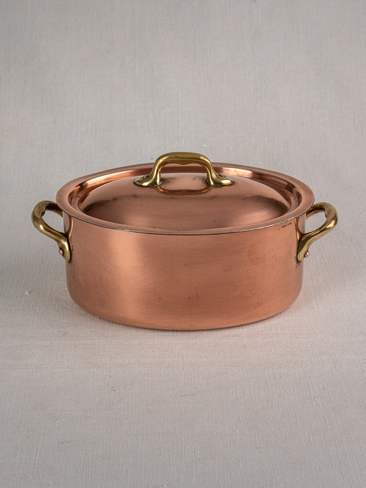 Oval vintage French copper cocotte with brass handles and domed lid on beige fabric background.