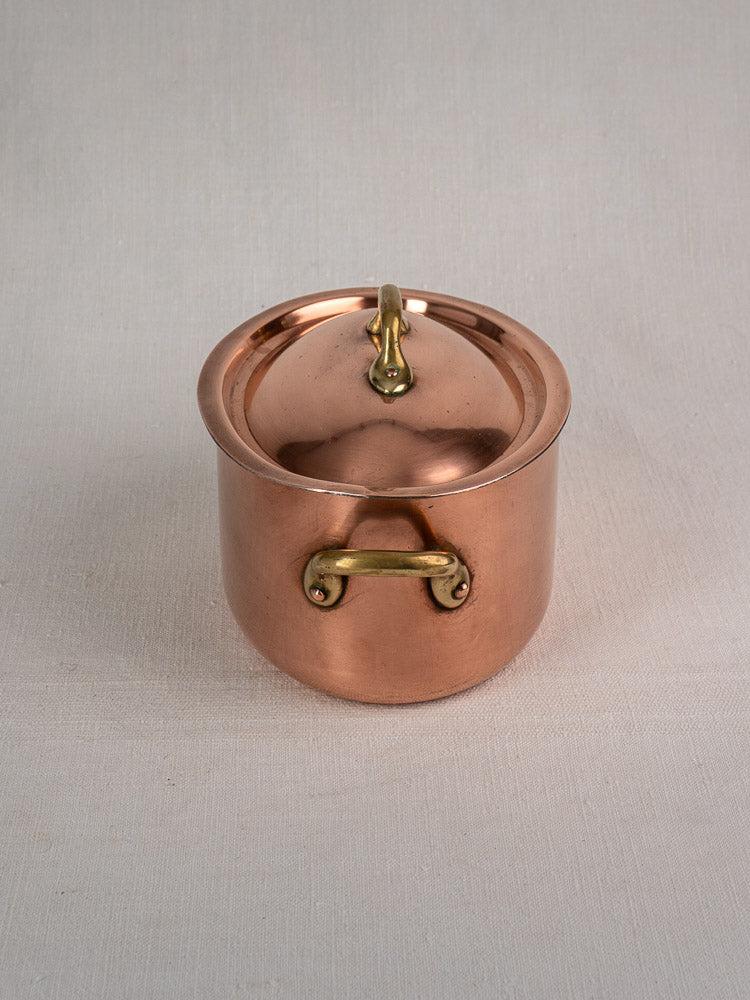 Vintage French copper cocotte with domed lid and brass handles on light fabric surface.