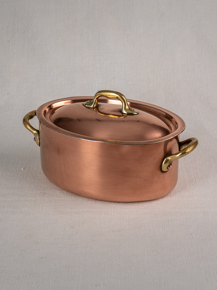 Vintage French copper cocotte with oval shape, brass handles, and domed lid on light textured surface.