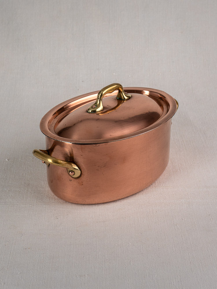 Vintage French copper cocotte, oval with domed lid and brass handles, on light beige textured surface.