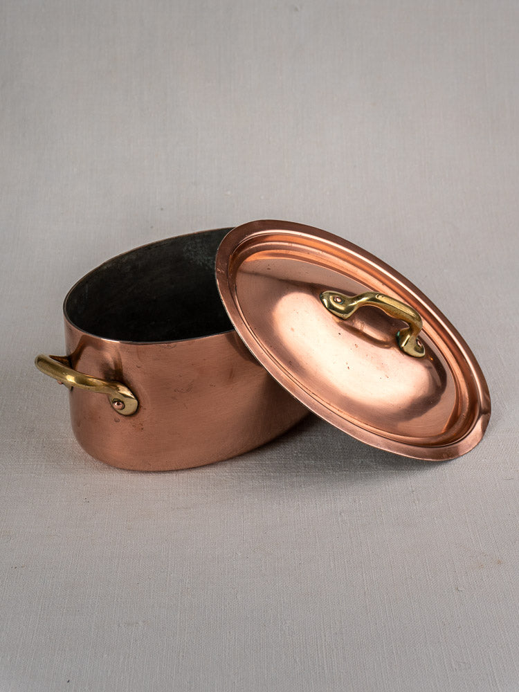 Vintage French copper cocotte with brass handles and domed lid beside oval pot on neutral background.