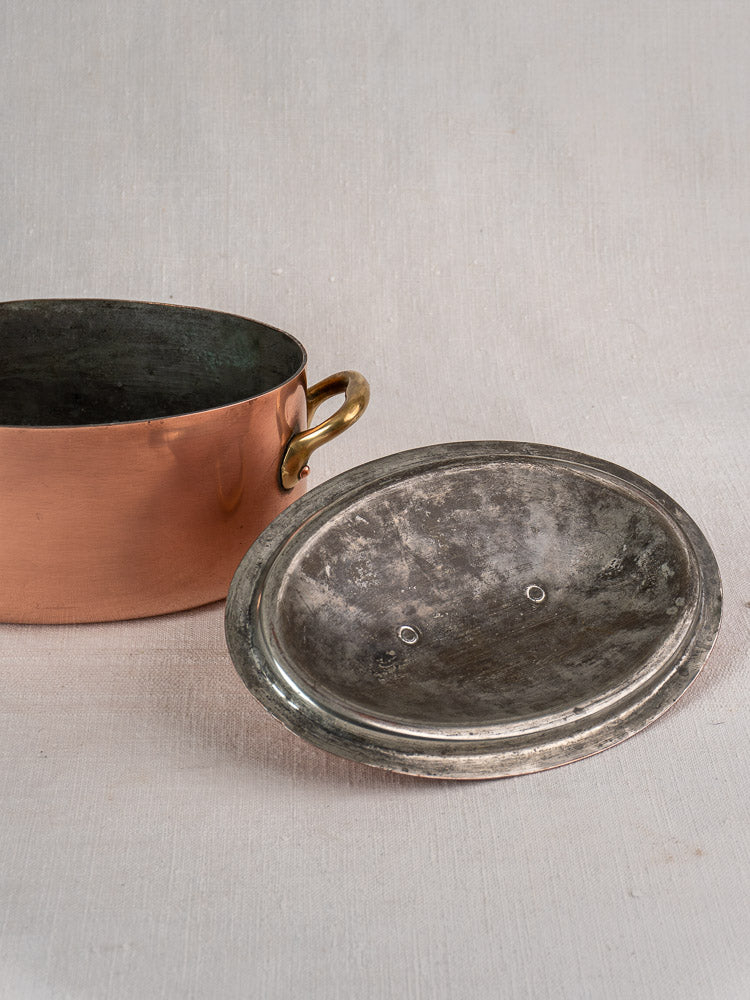 Vintage French copper cocotte with brass handles and domed lid on a light textured surface.