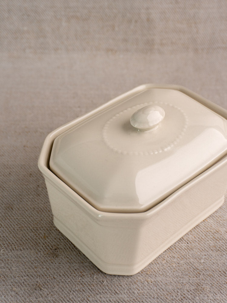 Antique French porcelain terrine, cream glazed, rectangular with lid and round knob, on textured beige fabric.