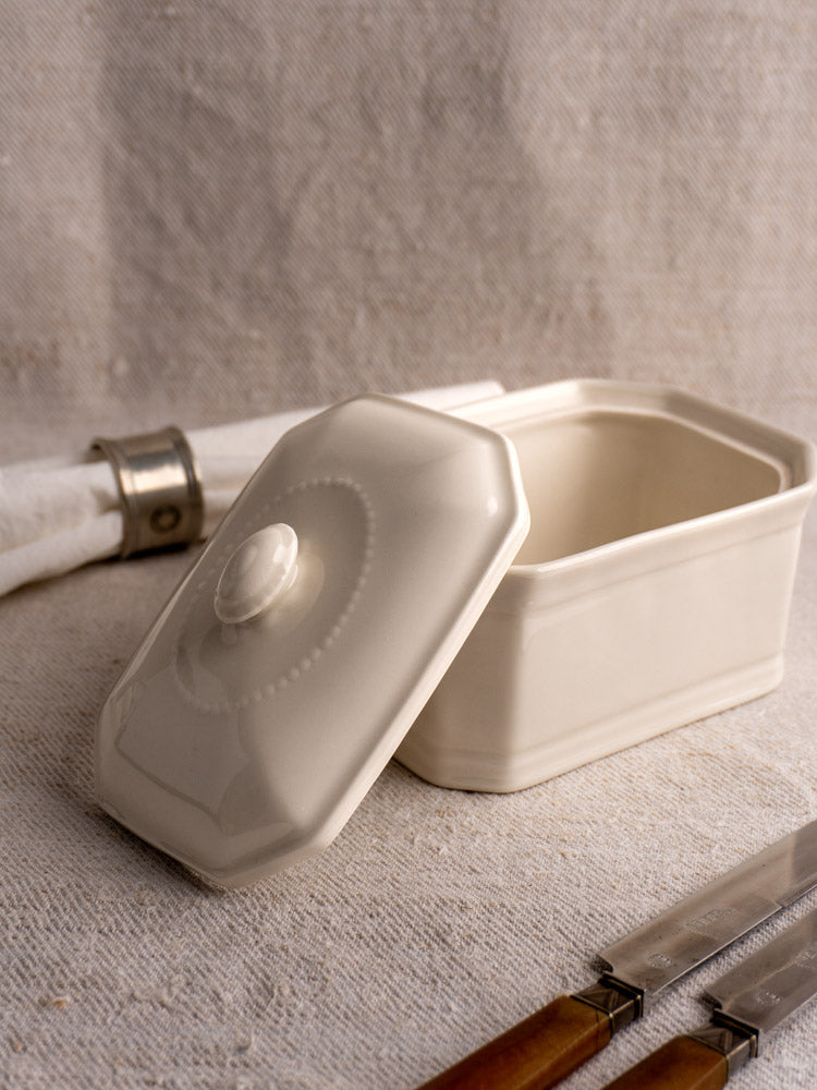 Antique French cream-glazed porcelain terrine with lid, displayed on textured fabric beside wooden-handled knives and napkin.