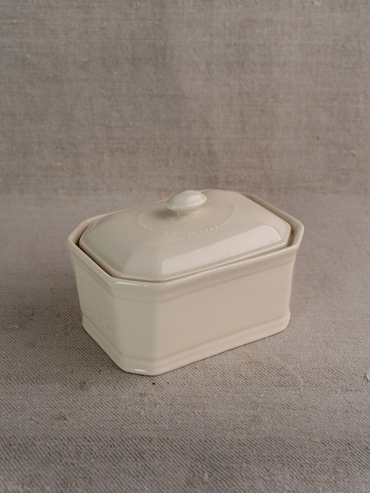 Small antique French porcelain terrine with cream glaze, hexagonal edges, embossed lid on beige textured fabric background.