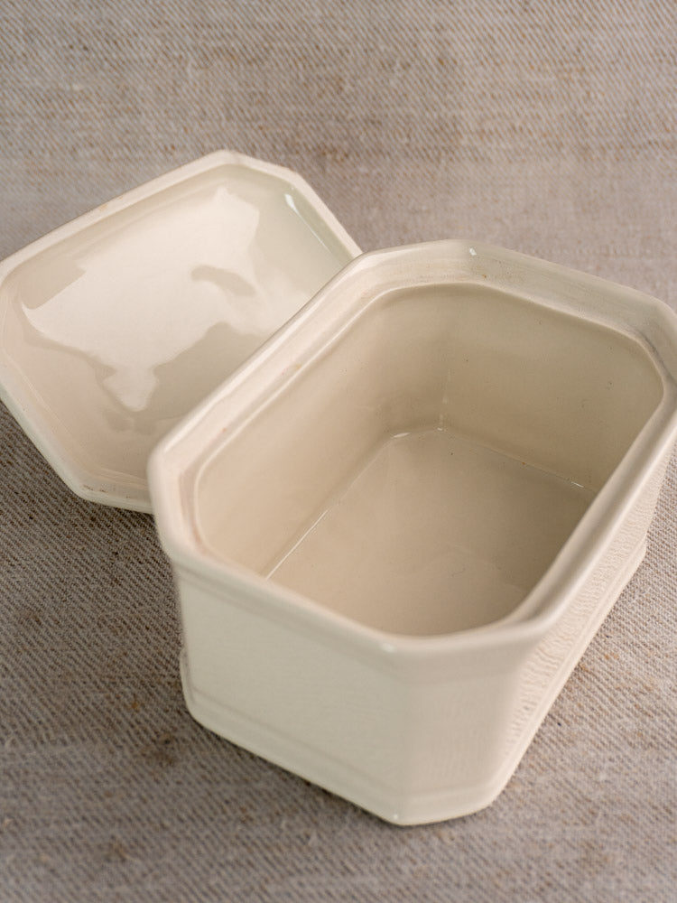 Antique French cream-glazed porcelain terrine with matching lid, rectangular shape, on beige fabric backdrop.