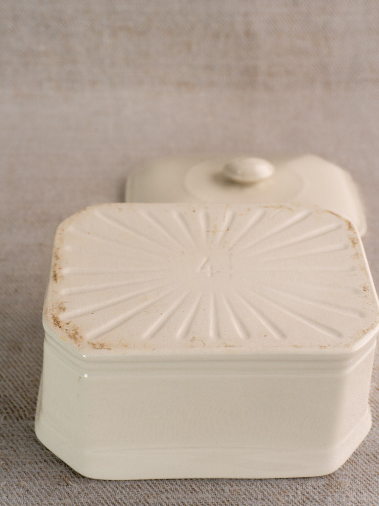Early 20th-century French porcelain terrine with cream glaze, sunburst lid, brown edge patina on neutral fabric surface.