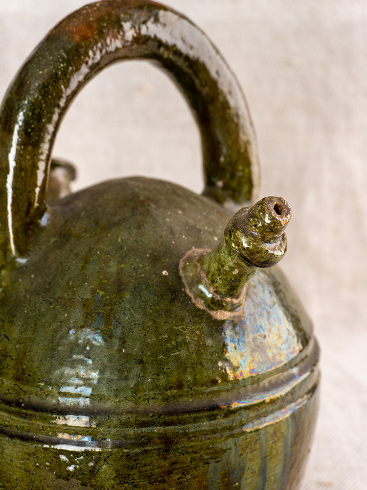 Antique French gargoulette, circa 1850, green glazed earthenware with rounded lid and curved handle on neutral textured background.