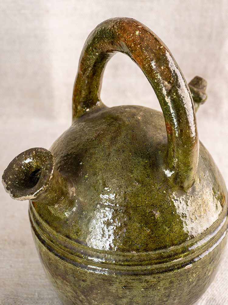 Antique French green-glazed earthenware gargoulette, rounded with curved handle and spout, on neutral fabric background.