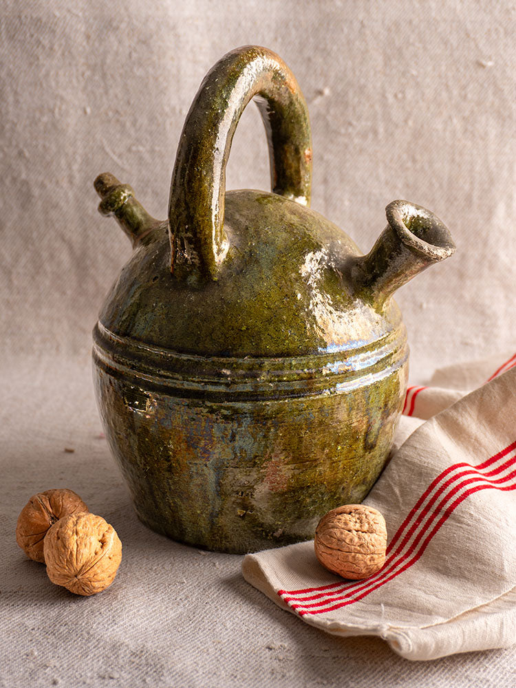 Antique French green glazed earthenware gargoulette, circa 1850, with two spouts on beige cloth near striped napkin and walnuts.
