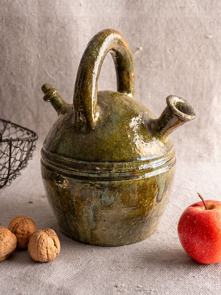 Antique French green glazed earthenware gargoulette jug with round body and two spouts on neutral fabric background.