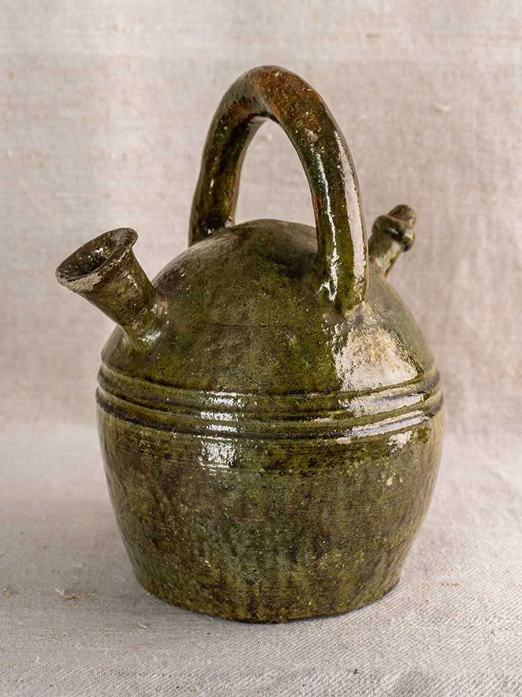 Antique French green glazed earthenware gargoulette jug with round body and two spouts on neutral fabric background.